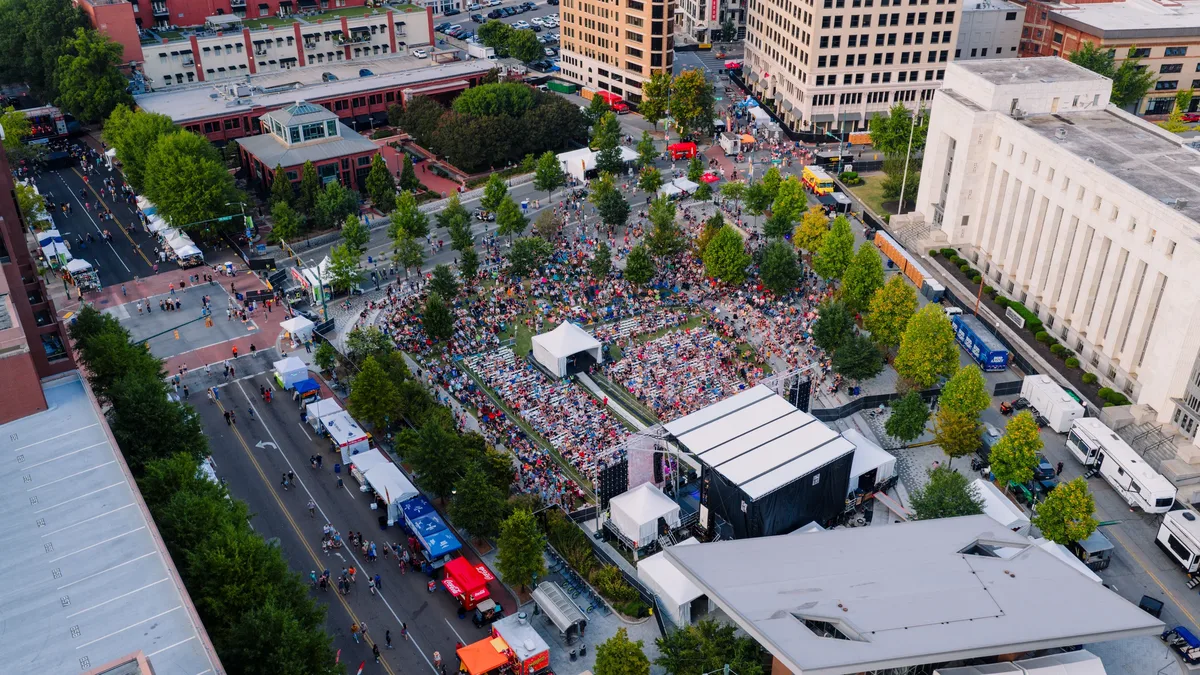 This aerial photograph shows a large crowd of people gathered in a city square, surrounded by trees and buildings, with a