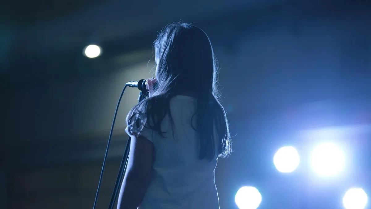 Music: A woman standing on a stage, facing away from the camera, with a microphone in front of her.