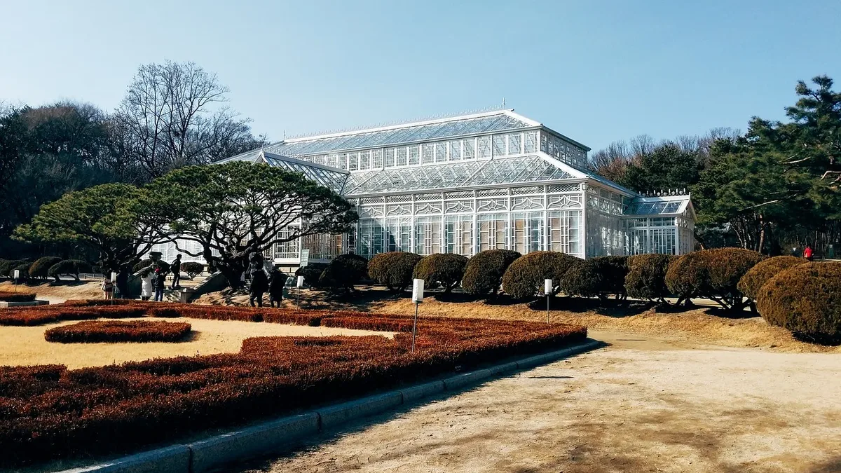 Plants: A large white building with a glass facade, surrounded by a well-manicured garden with trees, bushes, and a dirt path
