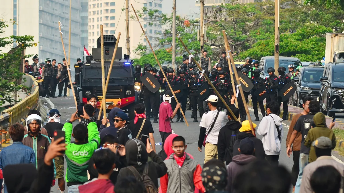 A crowd of people walking down a city street, with a line of police officers standing in front of them, holding wooden