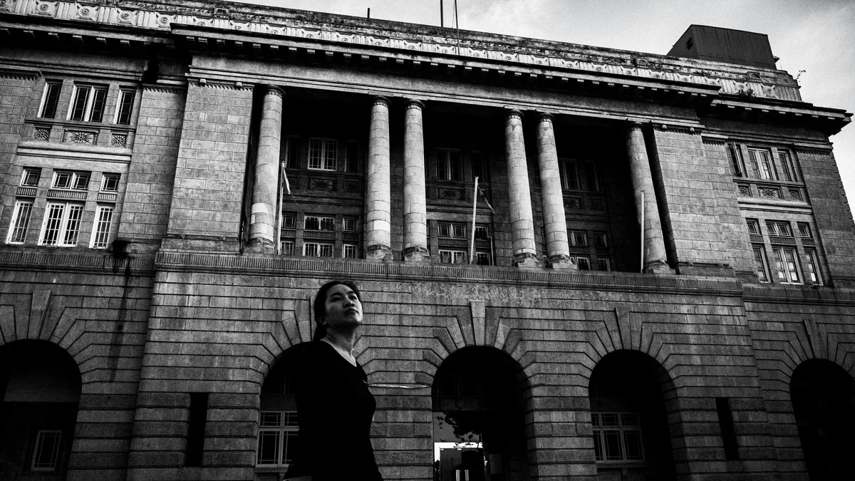 Implementation: A man standing in front of a large stone building with columns and arched windows.