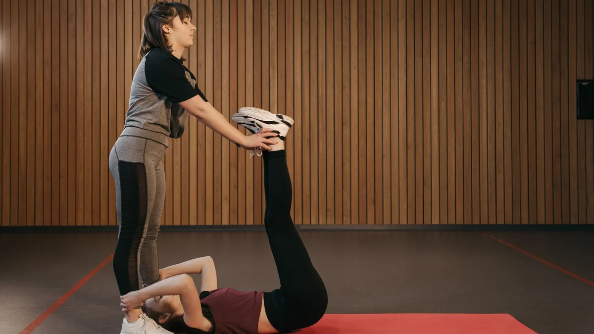 A woman holding a stack of folded white towels while standing over a woman lying on a red yoga mat, with the woman on the