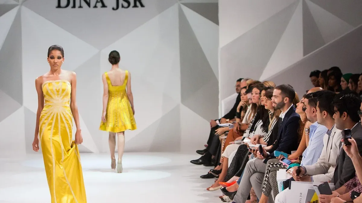 A runway fashion show with two models walking down the catwalk in yellow dresses, with an audience seated on either side