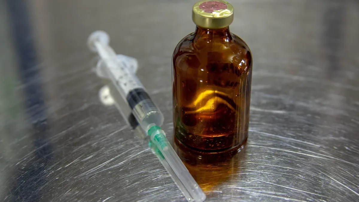 Money Cover: A small brown glass bottle with a gold cap and a syringe lying next to it on a metal surface.