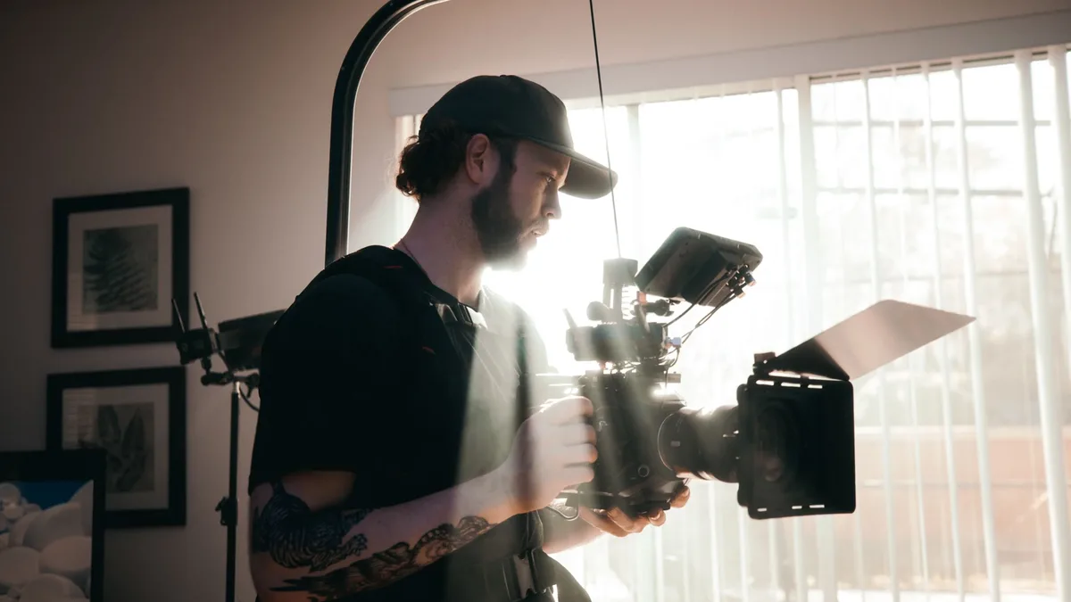 A bearded videographer in a black shirt operates a film camera inside a sunlit room, showcasing filmmaking in action