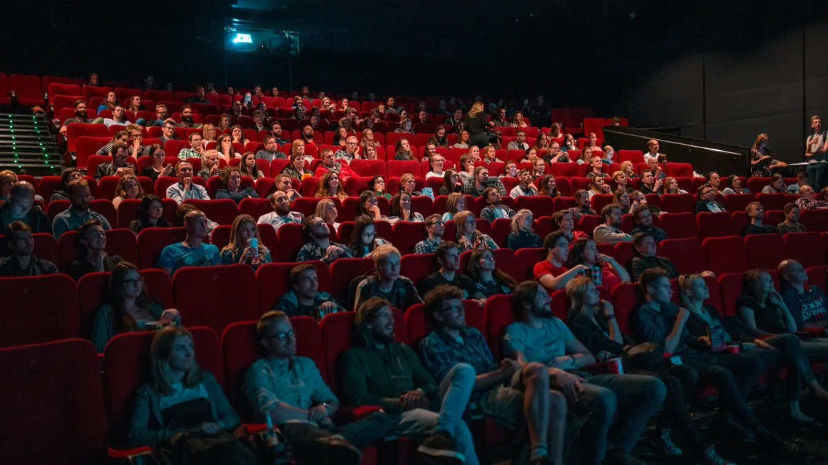 Box Office: A large group of people are seated in a dimly lit theater, watching a movie.