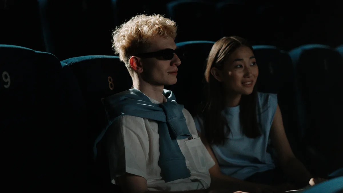 A man and woman are seated in a dimly lit movie theater, with the man wearing 3D glasses and a blue scarf, and the woman