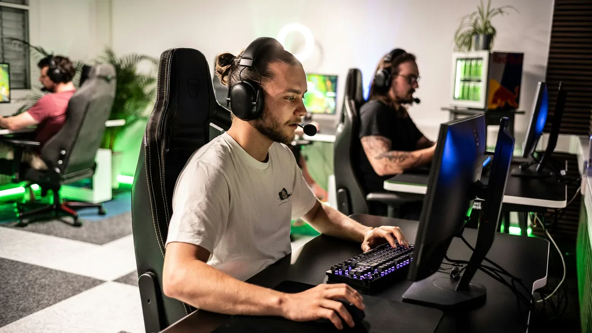 Teams: Three men are seated at computer stations, each wearing a headset and focused on their computer screens.