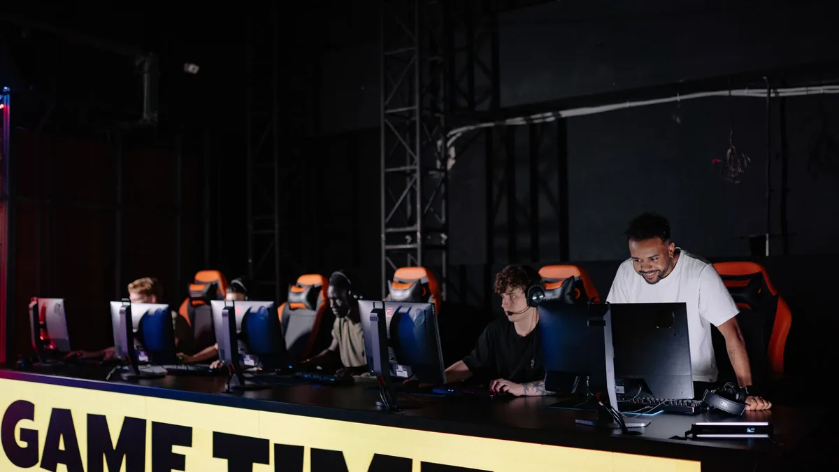 A group of people are seated at a long table with multiple computer monitors, with a large sign reading "GAME TIME" in