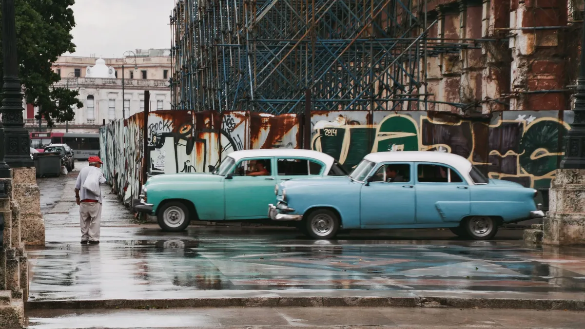 Marques: Two vintage cars parked on a wet street in front of a building with graffiti, with a person walking on the sidewalk.