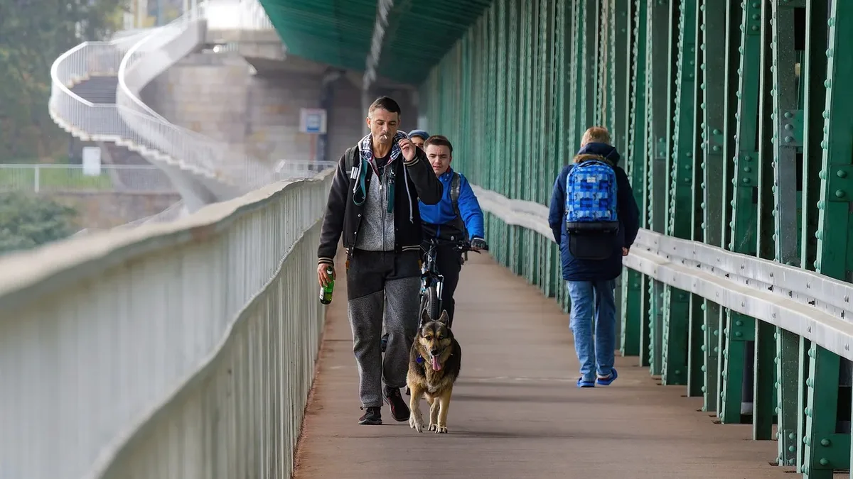 A man walking with a dog on a bridge, accompanied by two other individuals and a railing on the left side.