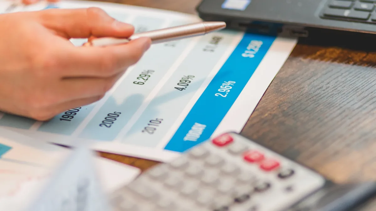 Inflation Currency: A person's hand holding a pen over a document with a calculator and laptop in the background.