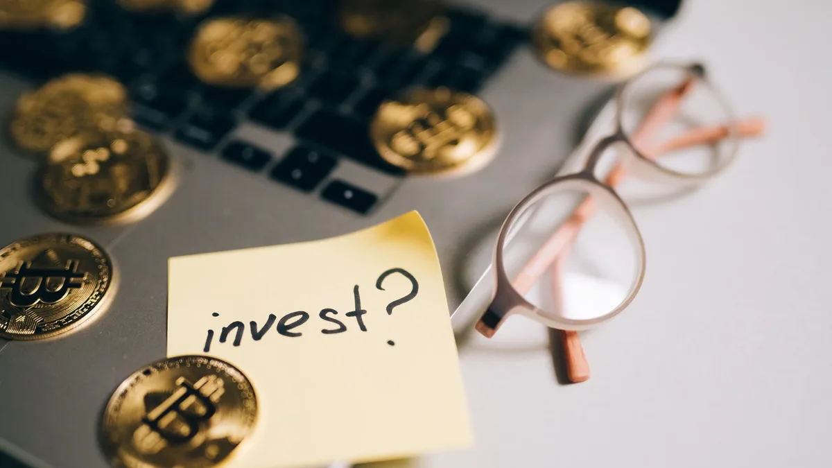 Getting Started: A close-up of bitcoin coins on a laptop with eyeglasses and a sticky note reading 'invest?'.