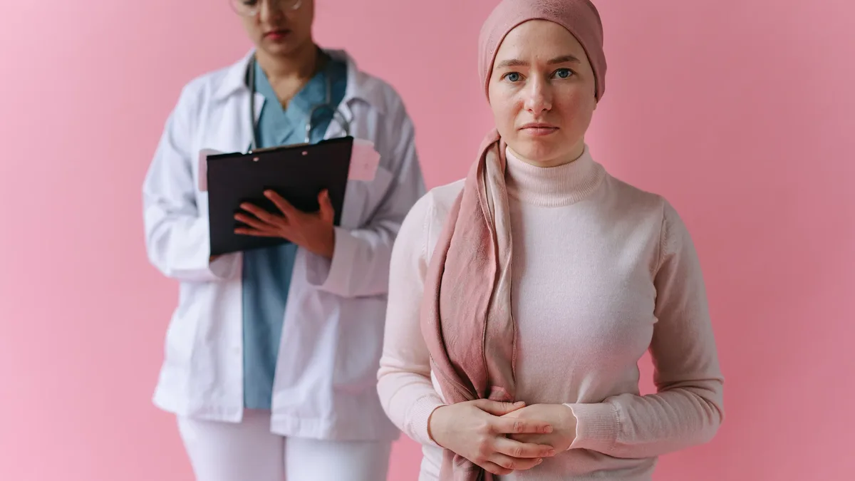 A woman wearing a pink headscarf and a light-colored turtleneck sweater stands in front of a pink wall, with a woman in a