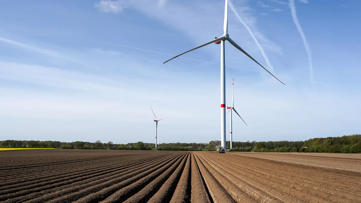 Impacts In Europe: A wind turbine standing in a field with rows of soil, set against a blue sky with clouds.