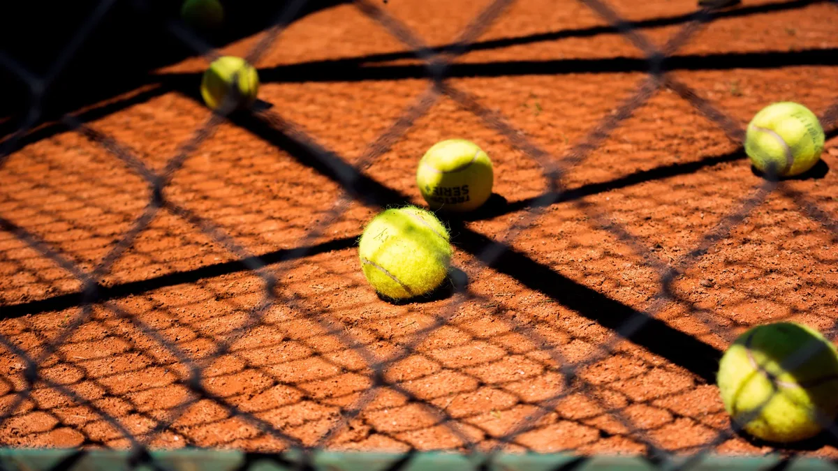 Players: Close-up view of tennis balls scattered on a clay court, captured through a chain-link fence.