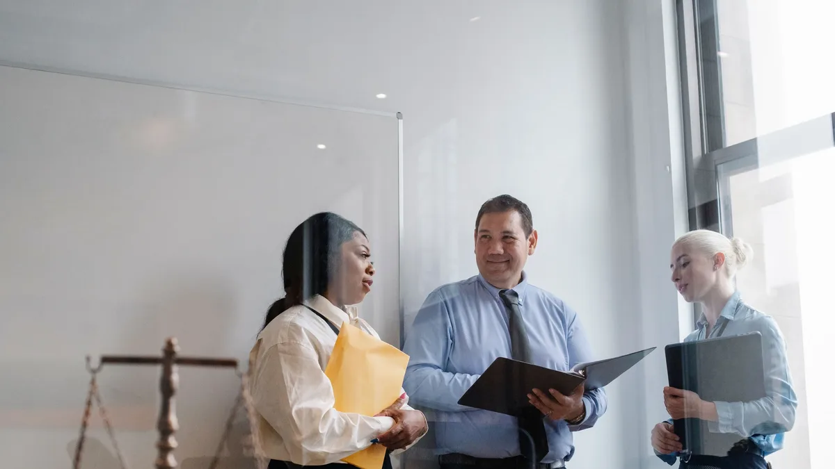 Stages: A man and two women standing in an office, with the man holding a black folder and the women holding yellow and black
