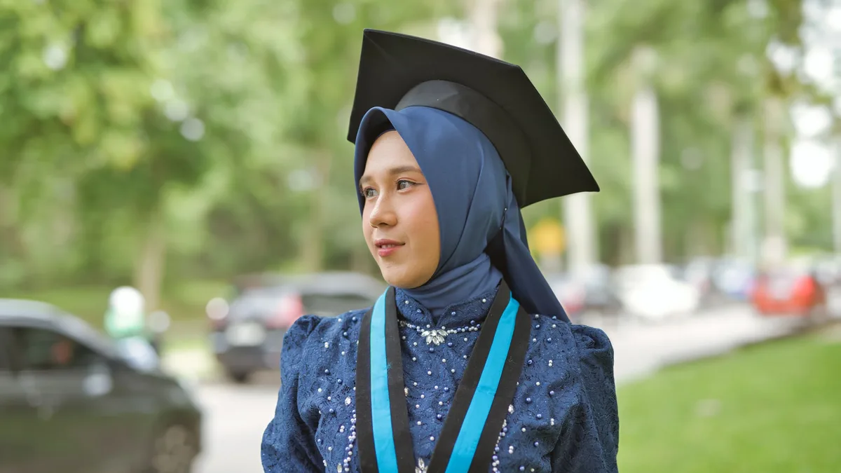By Level: Young woman in hijab wearing a graduation cap, celebrating achievement outdoors.