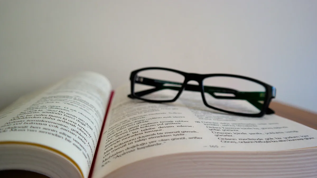 By Funder: A pair of black-framed glasses resting on an open book with a red bookmark, set against a white wall.