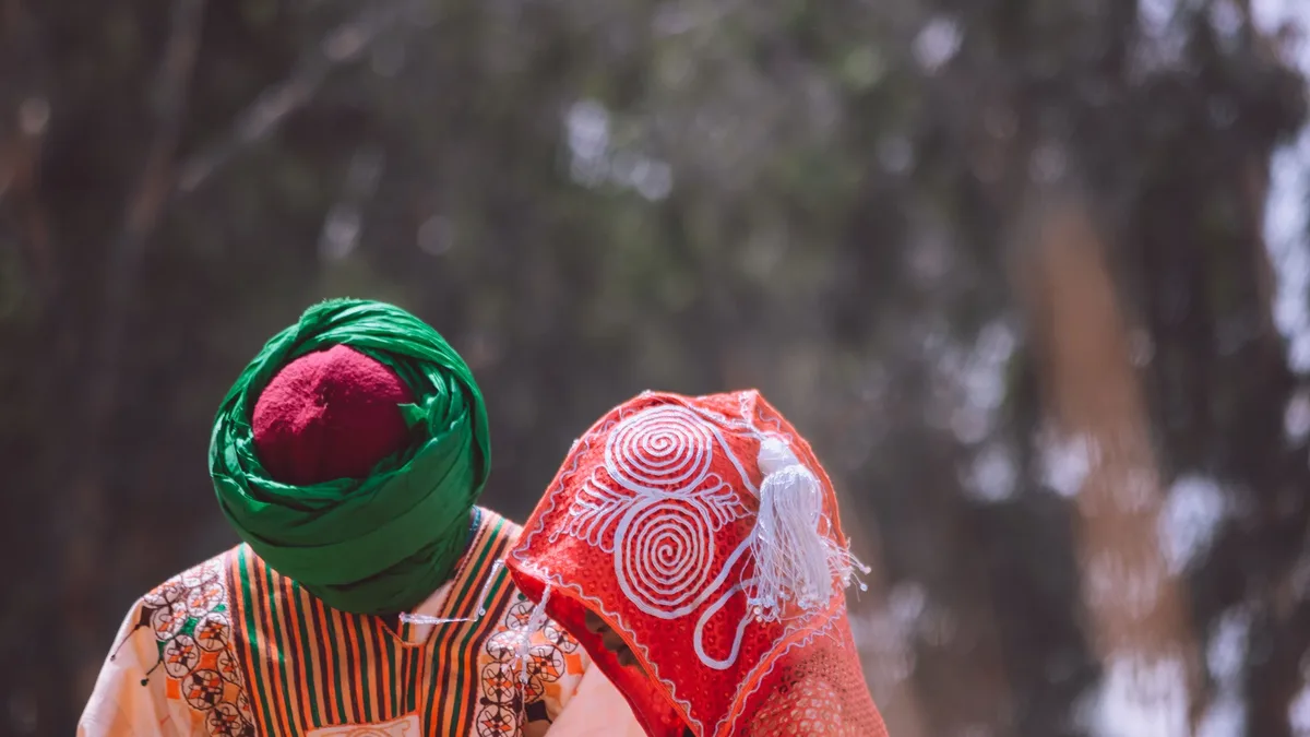 A person wearing a green turban and a red headscarf, with a white and red patterned scarf draped over their shoulder, set