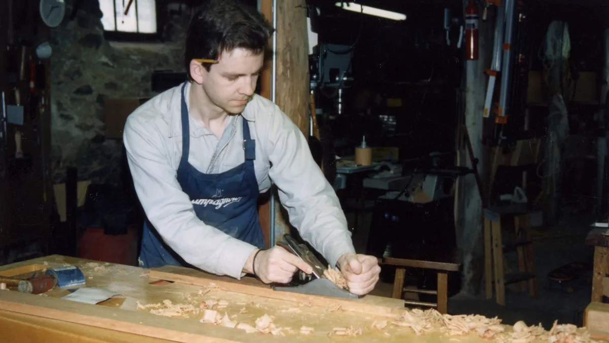 Pieces: A man is shown using a hand plane to shave wood shavings from a plank of wood on a workbench in a workshop.