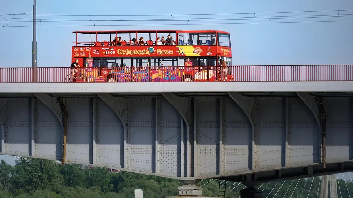 A red double-decker bus with people on board crossing a bridge over a body of water, with trees visible in the foreground