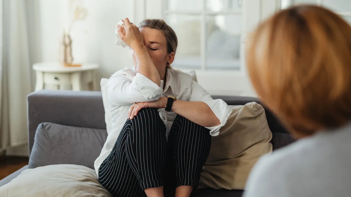 Systems: A woman wipes tears during a therapy session on a couch indoors.