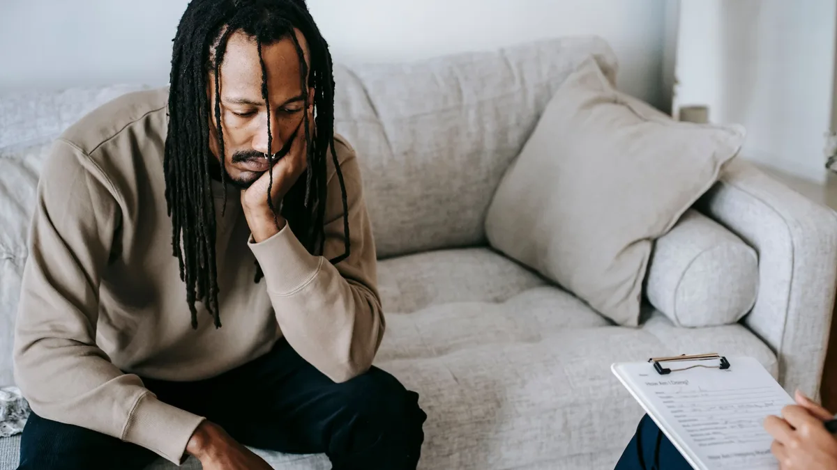 Upset African American male with dreadlocks leaning on hand and looking down while sitting on couch near crop psychologist