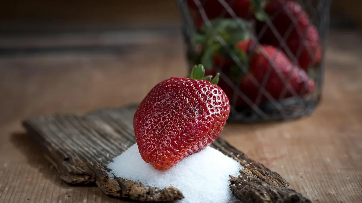 A strawberry sitting on a pile of white sugar, with a wire basket of strawberries in the background, all placed on a wooden