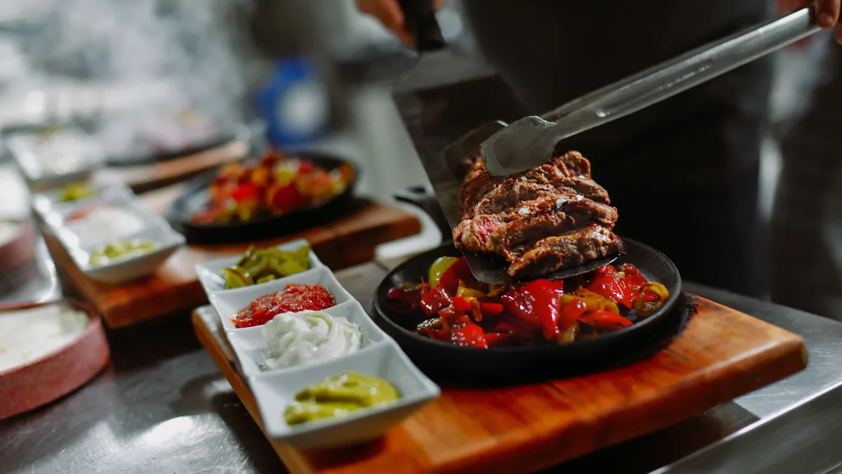 A person is preparing a meal consisting of sliced meat and vegetables on a sizzling skillet, accompanied by a selection of