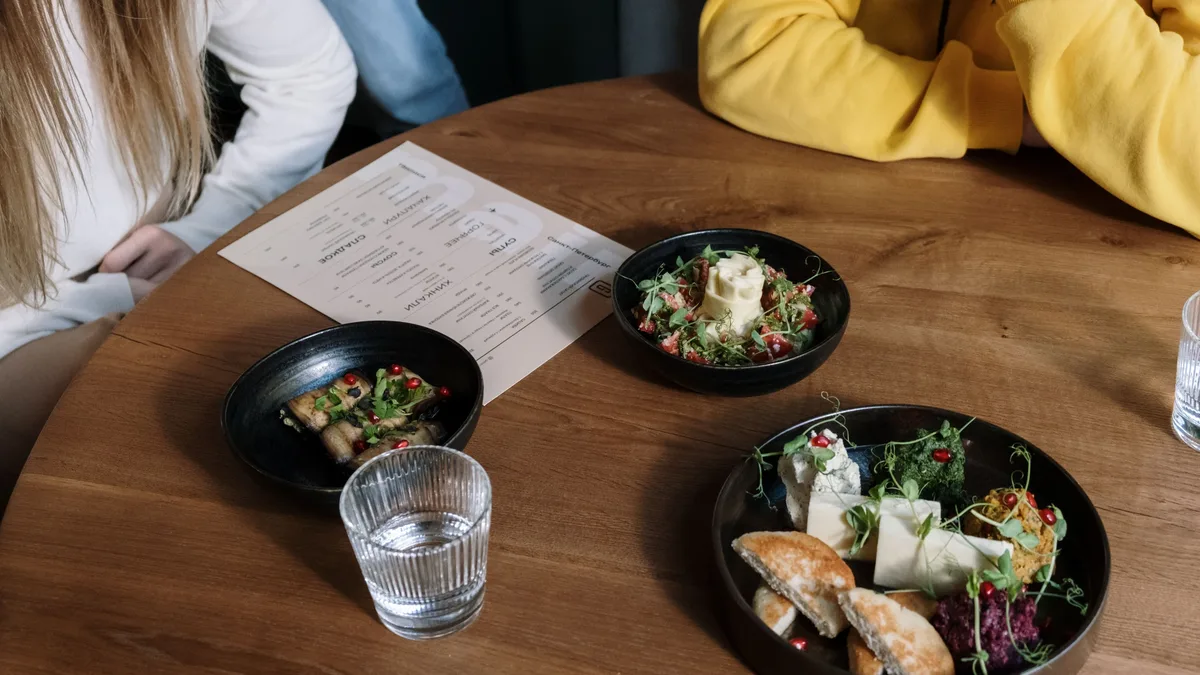 A round wooden table is set with three black bowls of food, a glass of water, and a menu, with two people sitting on either