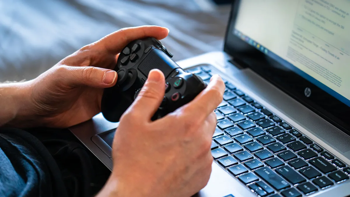 Reviews: Person holds a gaming controller while interacting with a laptop, emphasizing modern gaming technology.