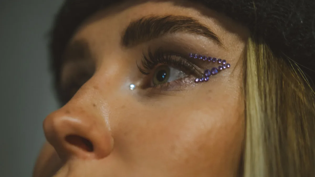Seasons: A detailed close-up of an eye with artistic makeup featuring purple beads and intricate eyeliner design.