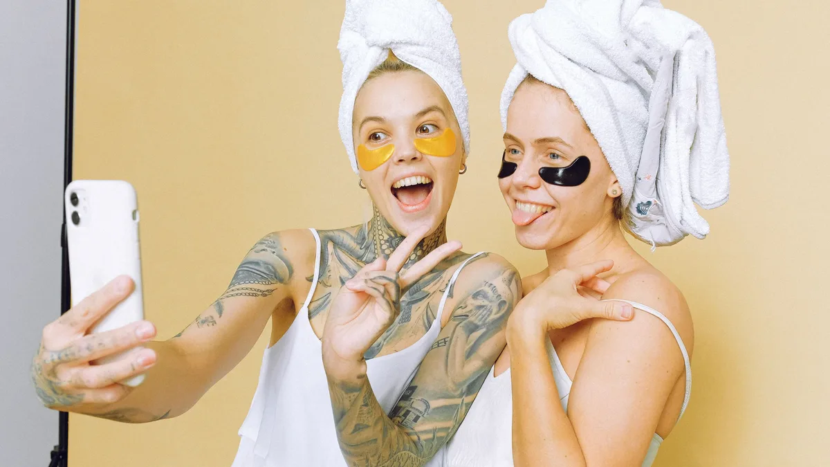 Two women enjoying a skincare routine with facial masks, taking a selfie wrapped in towels against a beige background