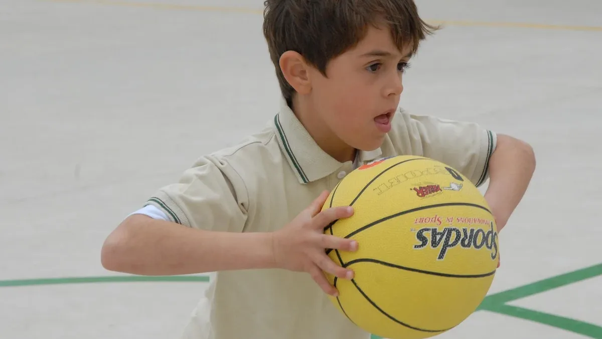 A young boy holding a yellow basketball with the word "Nordas" written on it, wearing a beige polo shirt with green stripes
