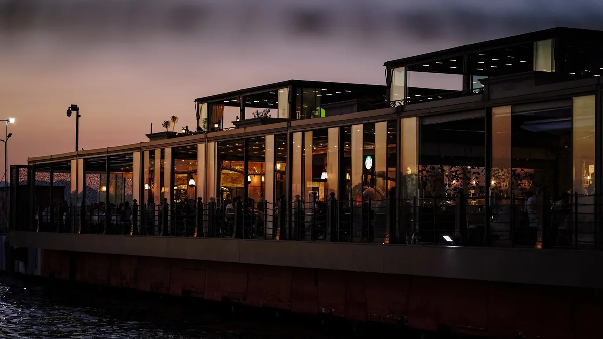 A modern restaurant by the water at twilight with warm interior lighting and sunset sky. Restaurants & Dining background