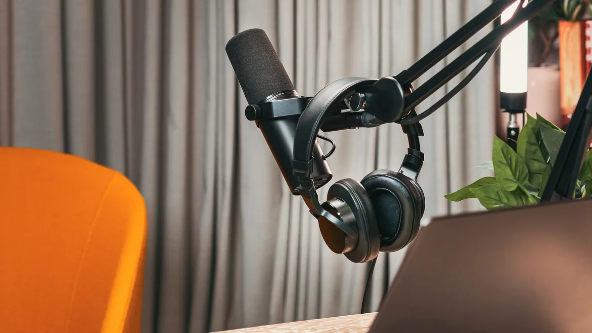 Formats: A laptop, headphones, microphone, and cell phone on a table in front of a chair and curtains.