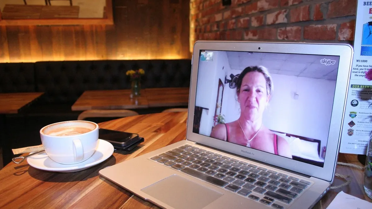 Cocina: A laptop on a wooden table with a woman's face on the screen, a cup of coffee, and a brick wall in the background.