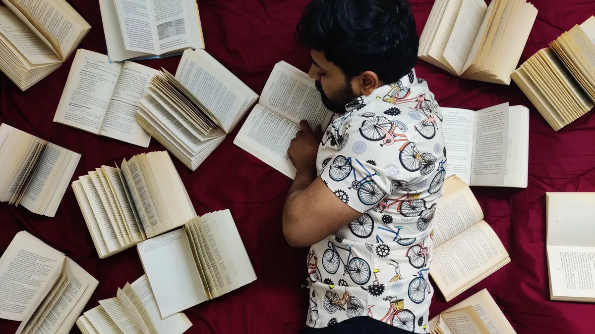 A man lies down reading on a red bedspread surrounded by open books, creating an artistic literary scene.