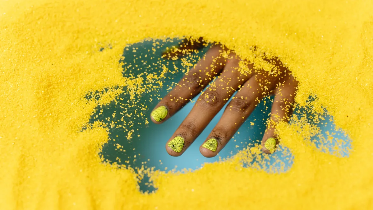 Nail Care: Close-up of a hand with creative nail art partially buried in vibrant yellow sand. Nails professional photography