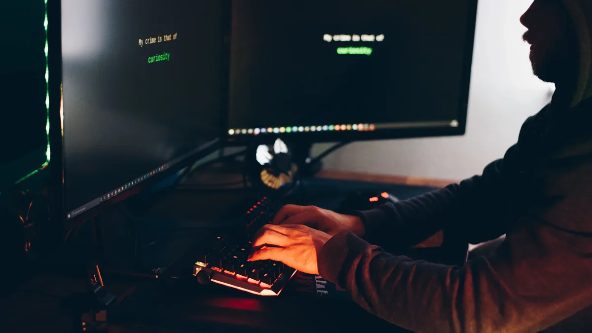 Side view of crop anonymous male cyber thief accessing information on desktop computer screens at dusk. Cybersecurity close